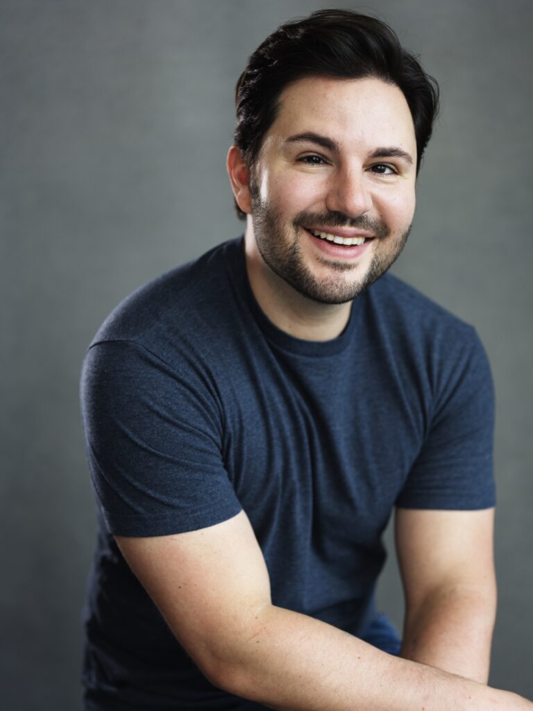 Smiling man with dark hair and light stubble in a navy T-shirt, posed against a gray backdrop."